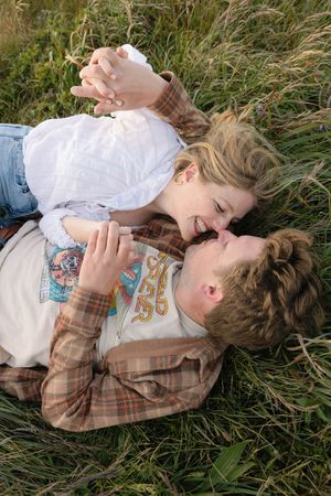 A romantic couple lying in tall grass wearing plaid and white clothing share an intimate moment together.