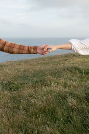 Hands reaching toward each other across grassy seaside cliff.