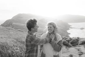 Black and white portrait of couple laughing together on coastal hillside.