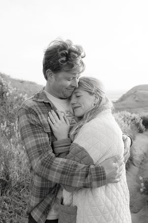 A tender black and white photo of a couple embracing outdoors in casual clothing against a mountain backdrop.