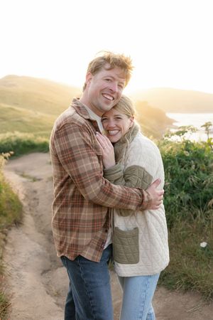 Happy couple embracing on a coastal path during golden hour with ocean views in background.