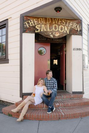 A couple sits on the brick steps outside The Old Western Saloon, a rustic bar entrance with vintage signage.