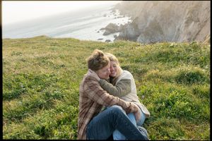 A romantic couple embraces in a grassy field overlooking coastal cliffs on a cloudy day.