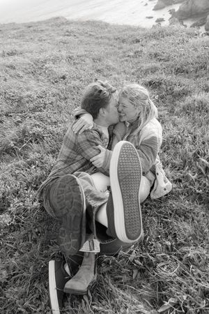 A black and white photograph shows a couple sitting close together on a grassy hillside wearing casual clothes and sneakers.