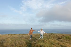 Two people walking hand in hand through a grassy hilltop meadow with ocean views and cloudy skies on the horizon.