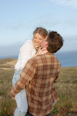 A joyful couple share a playful moment on a coastal clifftop with the ocean in the background.
