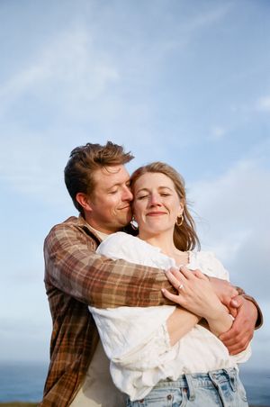 A couple shares playful embraces against a bright blue sky and ocean backdrop.