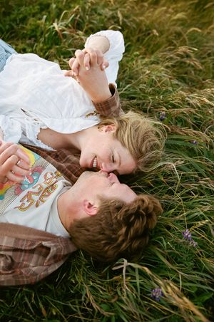 A couple lies in tall grass, laughing and embracing in casual vintage attire.