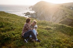 Two people sitting together on a grassy coastal cliff overlooking dramatic ocean views at golden hour.