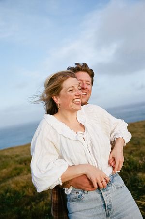 Candid moment of couple laughing together on breezy coastal cliff with ocean view.