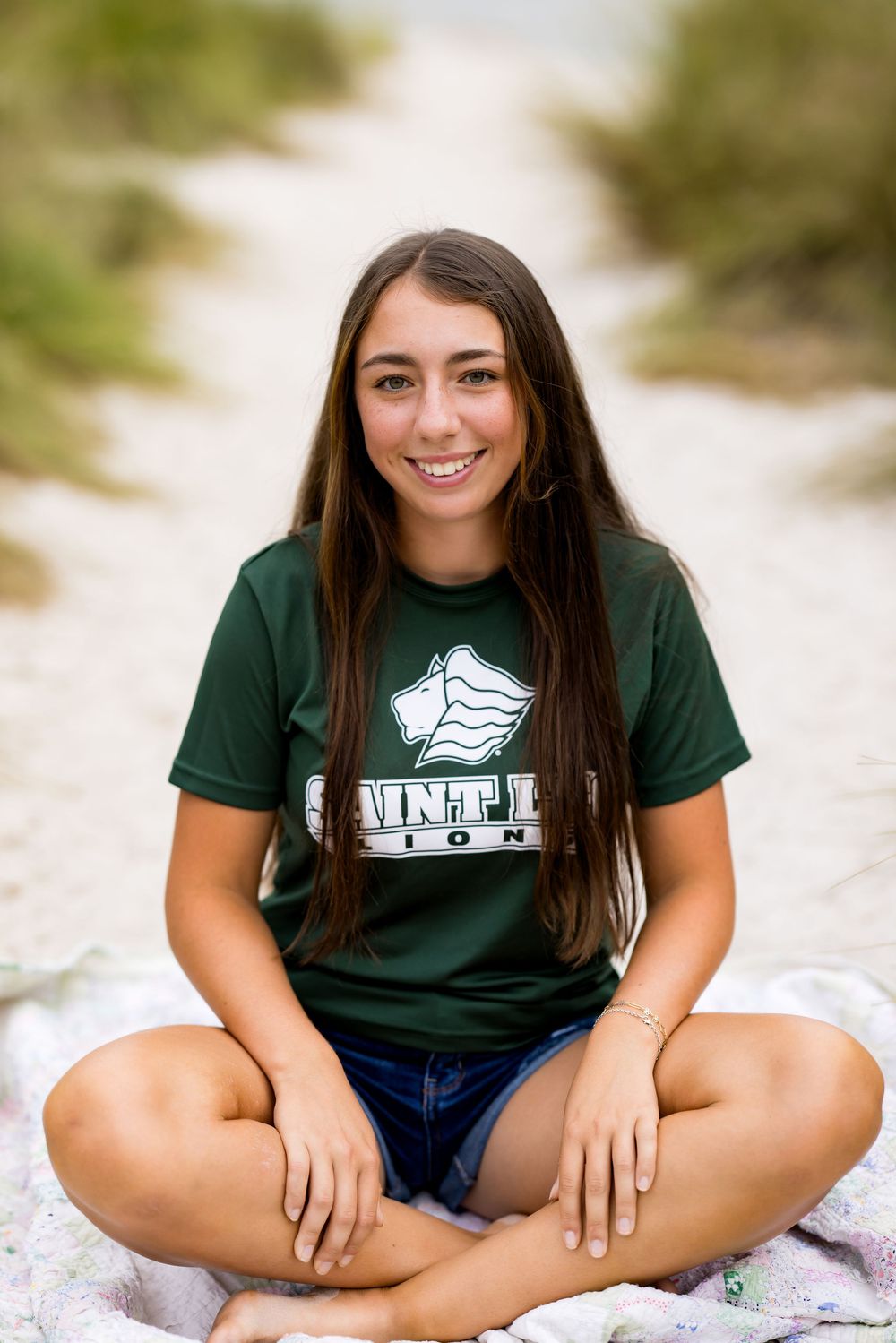 Sitting cross-legged on blanket wearing green t-shirt and denim shorts outdoors.