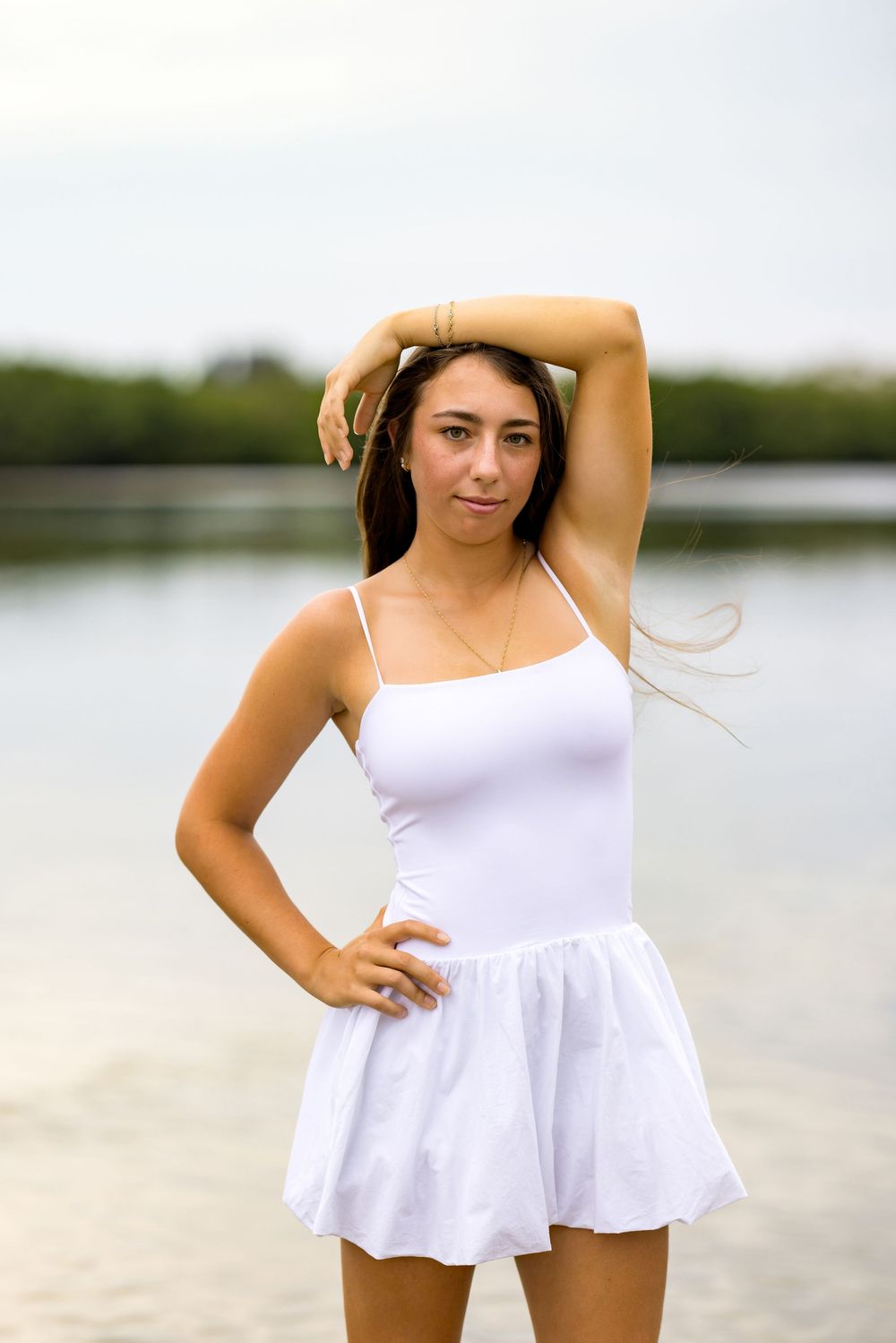 Posing in flowing white sundress by waterfront with arm raised.