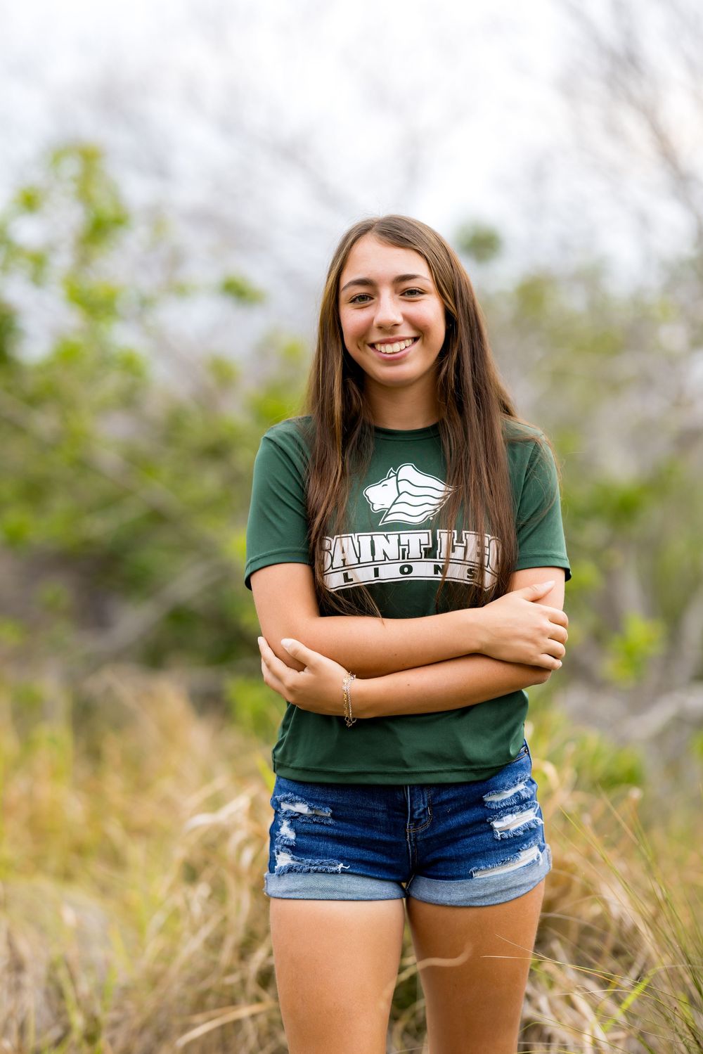 Outdoor portrait of someone in a green Point Park t-shirt and denim shorts standing with crossed arms.