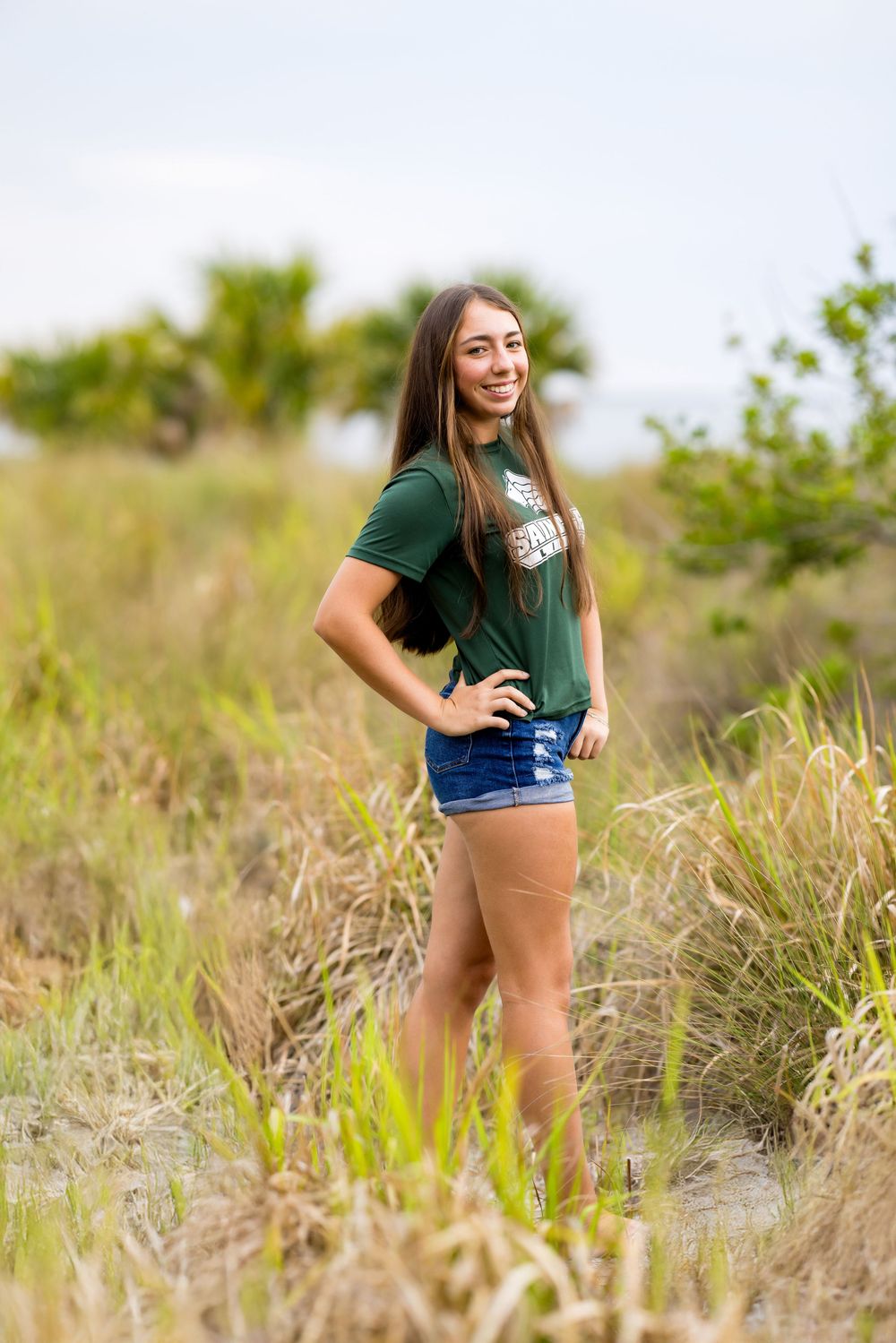 Woman in green t-shirt and denim shorts poses in grassy coastal landscape.