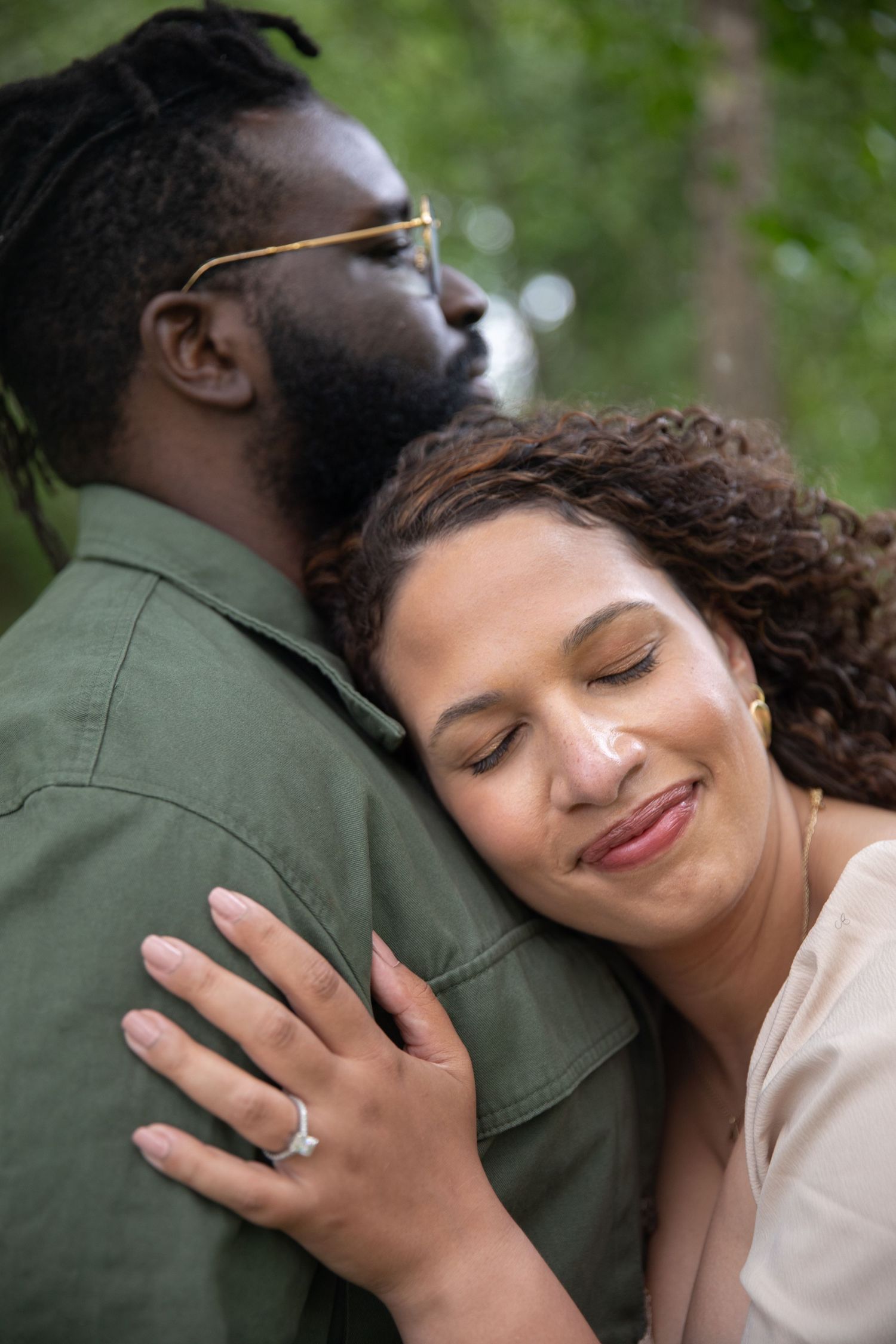 A couple sharing a tender moment outdoors as the person in white displays an engagement ring on their hand.