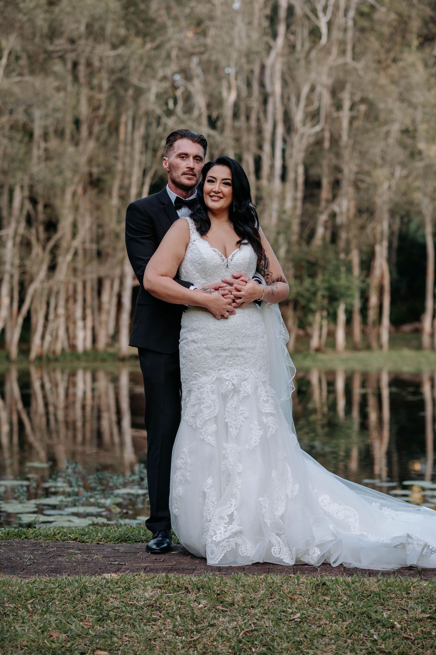 Newlyweds pose together by a serene pond with trees in the background as the bride wears a lace mermaid wedding gown.