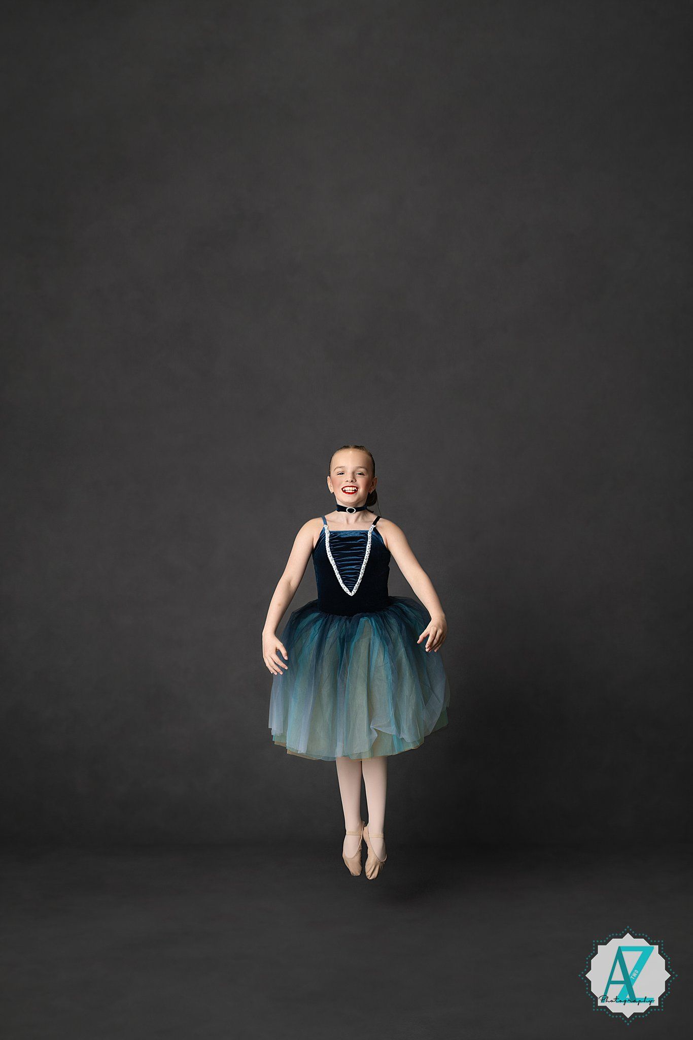 Young ballet dancer in turquoise ombre tutu dress and pearl necklace leaping gracefully against dark studio backdrop.