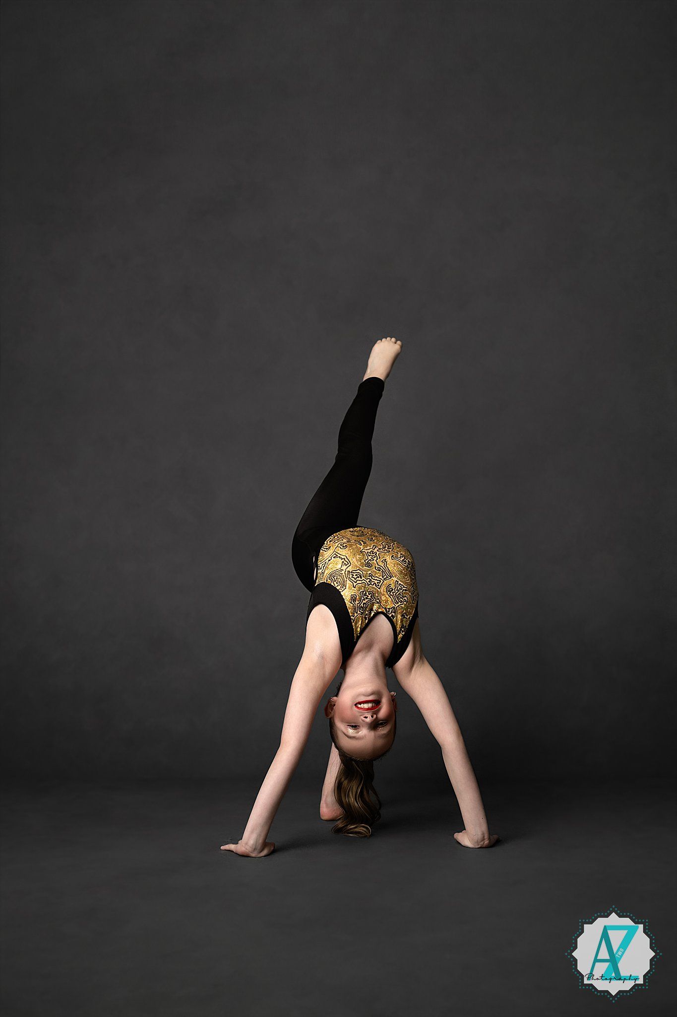 A gymnast in a gold and black leotard performs a split handstand against a dark studio backdrop.