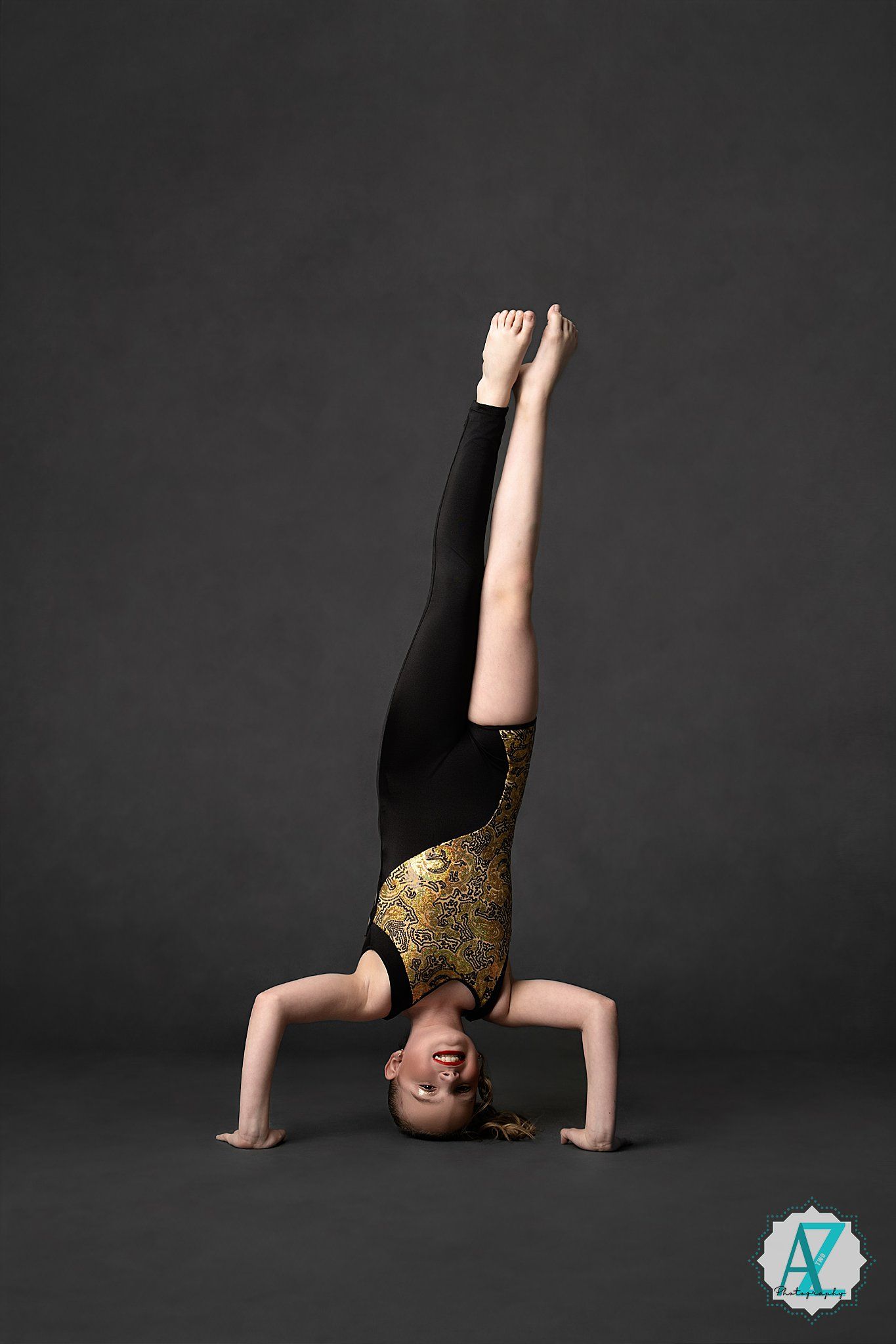 Gymnast performing a headstand split in a black and gold leotard against dark background during artistic photography session.