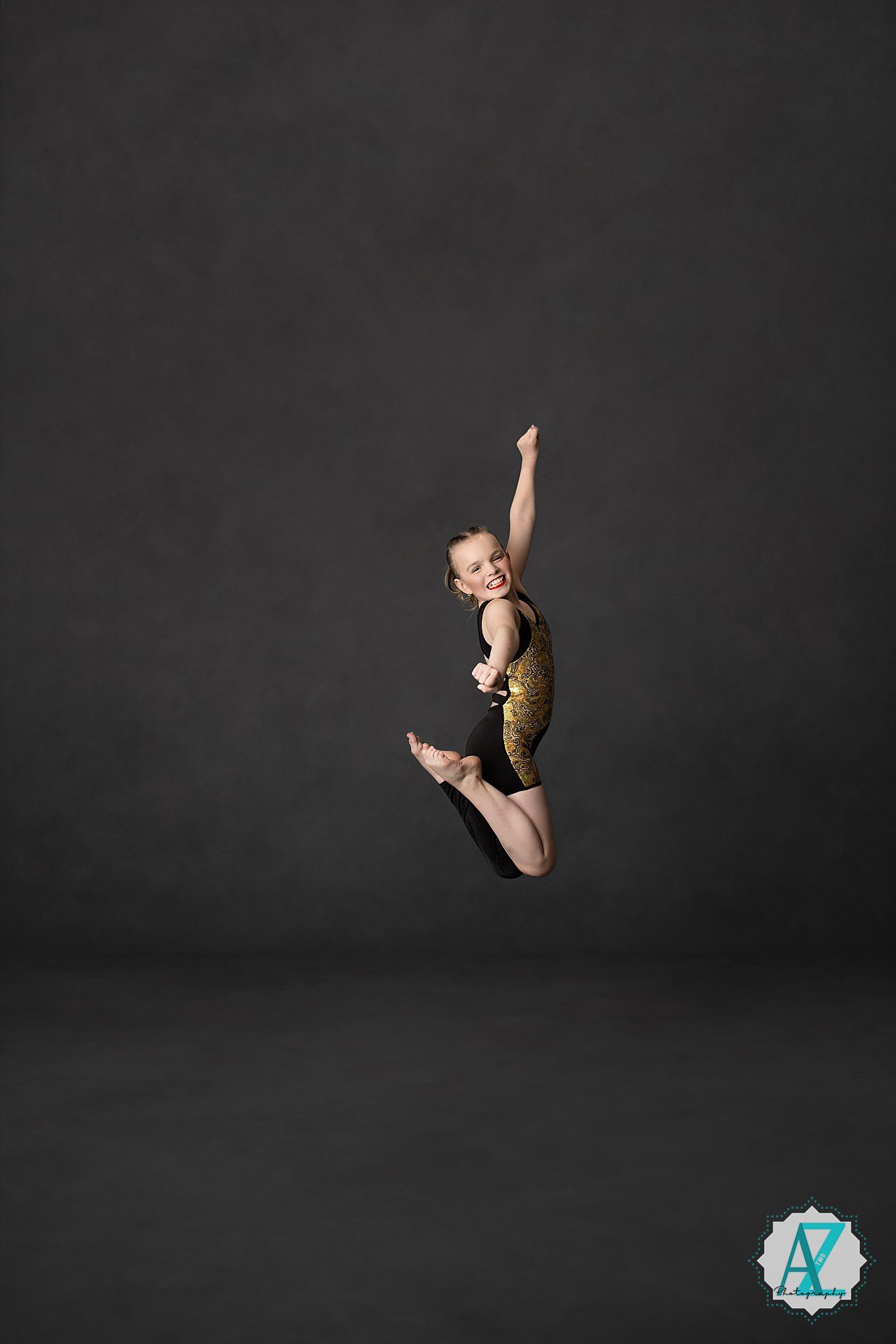 Gymnast in gold leotard performs graceful mid-air jump pose against dark studio backdrop.