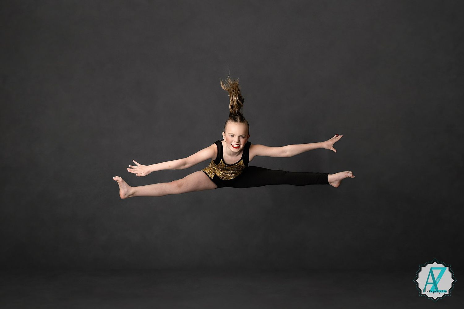 A dancer performs a split leap in the air against a dark backdrop wearing a black dance outfit.