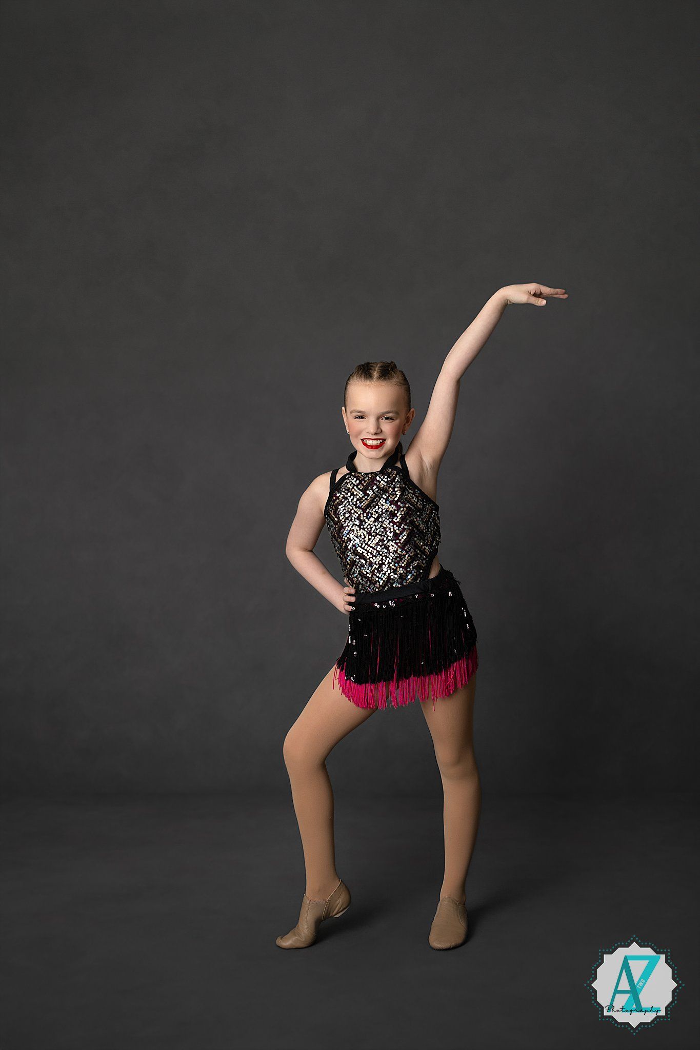 A dancer in a black sequined costume with pink fringe strikes a dynamic pose against a dark gray backdrop.