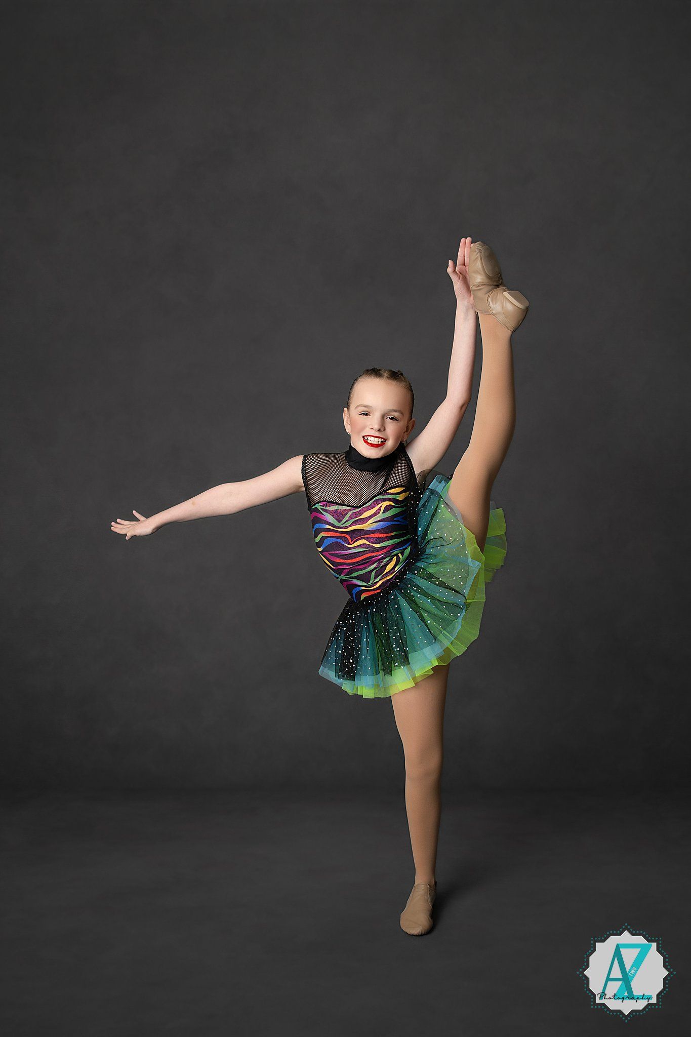 Young dancer in colorful leotard and tutu performs graceful high kick pose against dark studio background.