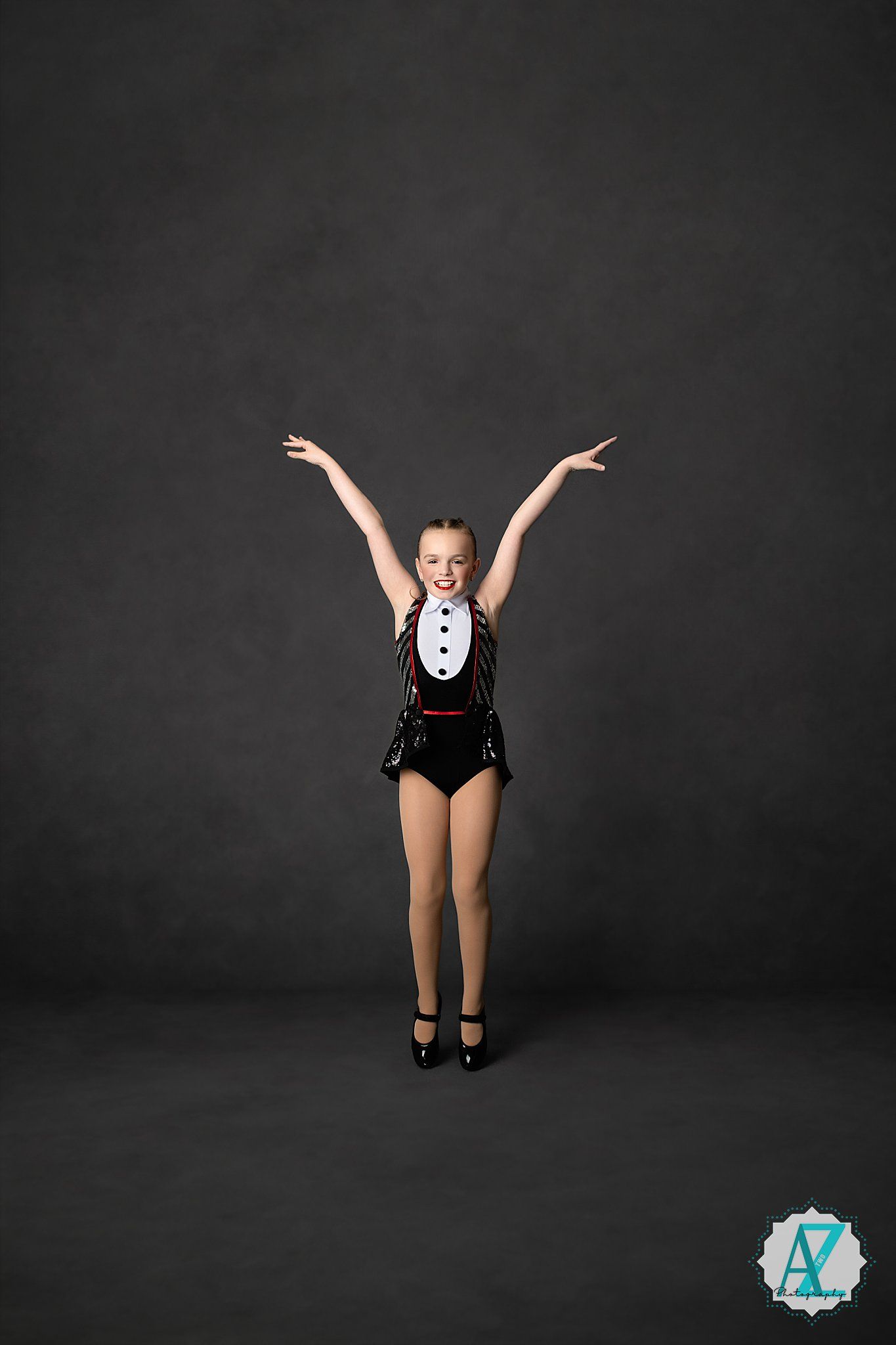 Young dancer in black leotard performs joyful jumps in a dark studio setting.