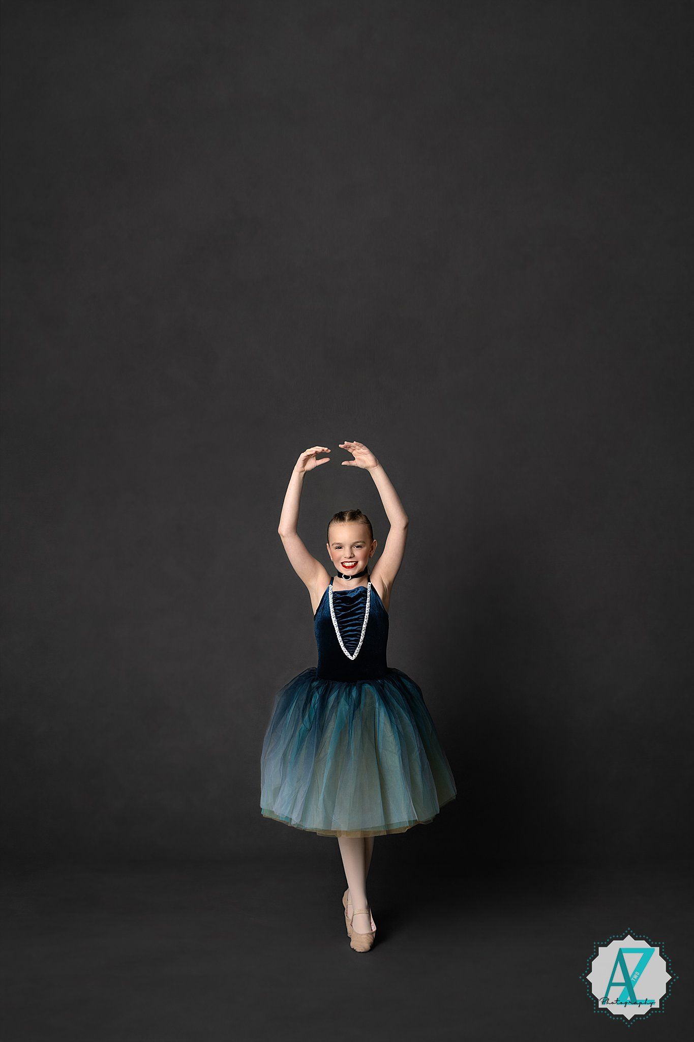 Elegant ballerina in a teal ombre tutu and pearls poses gracefully on pointe against dark backdrop.