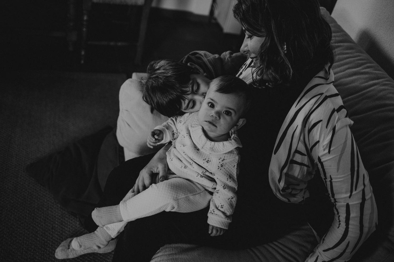 Mãe com a bebé no colo ganhando beijo do irmão mais velho  sentada no sofá da casa em uma foto preto e branco, sessão de família em Portugal
