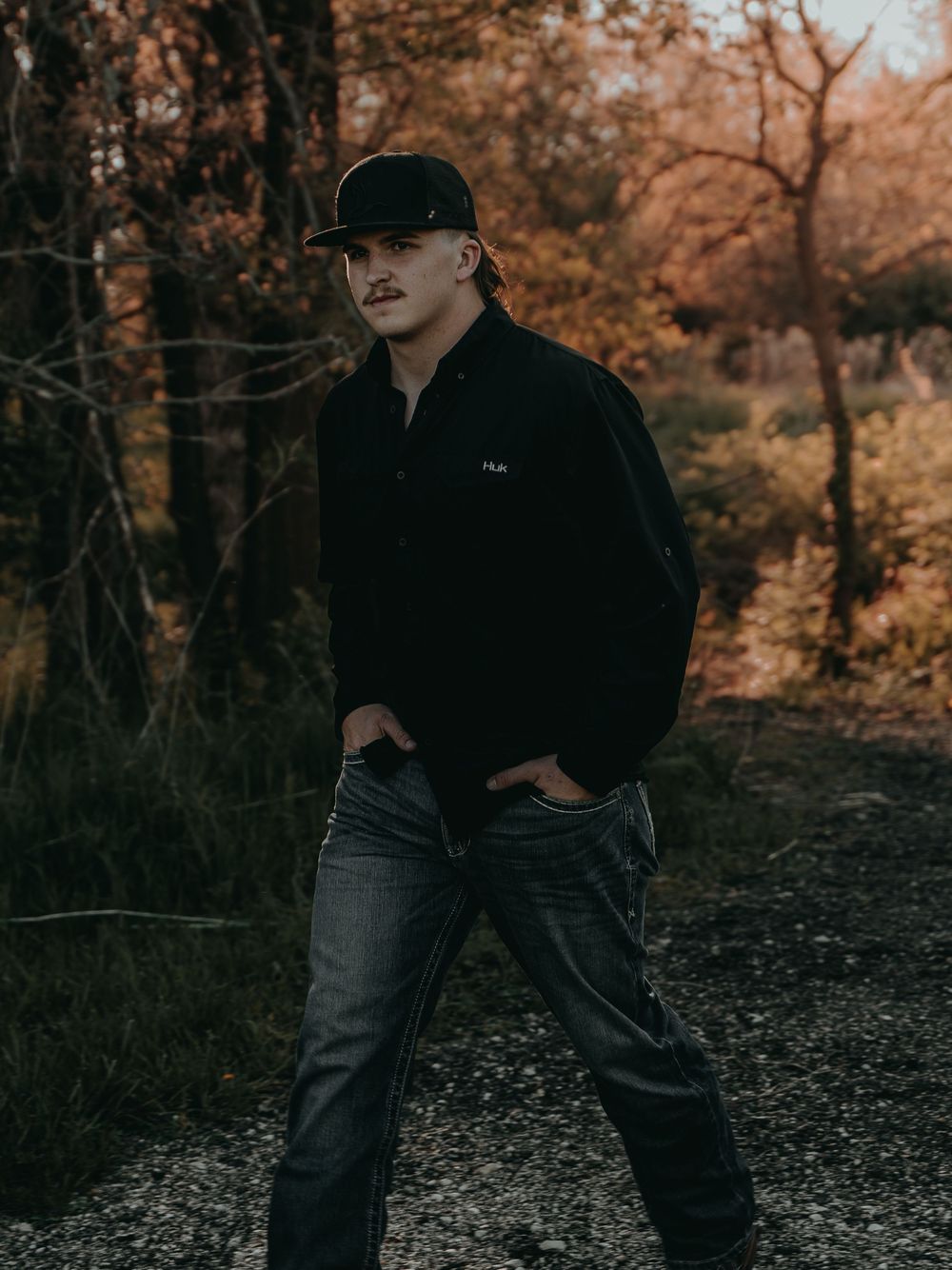 Outdoor Senior Portrait with autumn foliage background showing senior in dark jacket and baseball cap on dirt path - Warsaw, Indiana