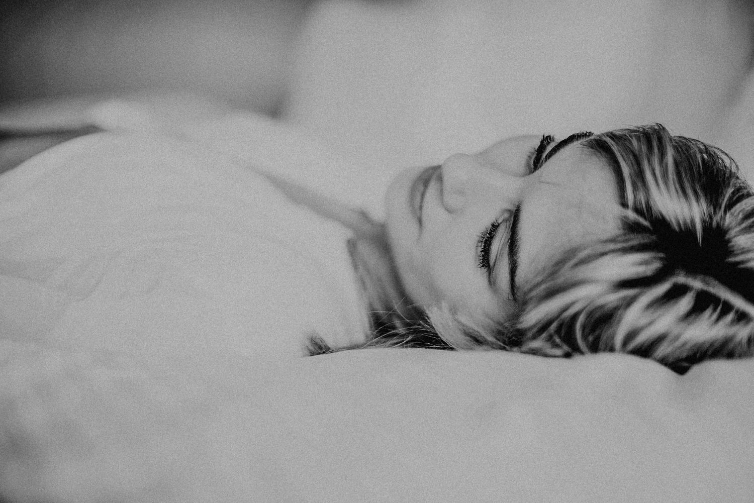 Schwarz-weiß-Foto einer Person, die entspannt auf einem Bett liegt. Die Person hat helle Haare und trägt ein helles Oberteil.