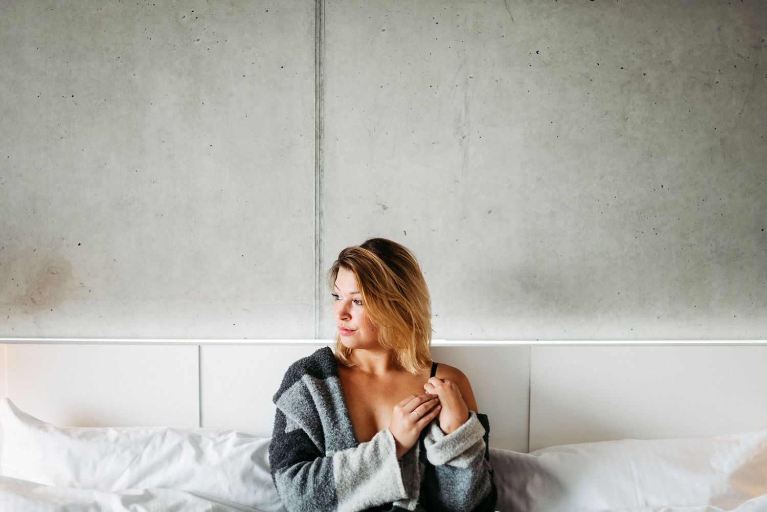 Frau mit blondem Haar sitzt auf einem Bett vor einer grauen Wand, trägt einen grauen Pullover und schaut nach links.