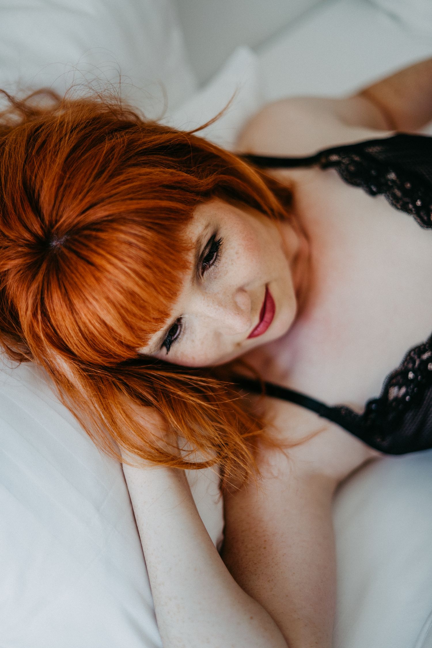 Eine Frau mit feurig roten Haaren posiert auf einem Bett. Sie trägt schwarze Unterwäsche und schaut sinnlich zur Seite.