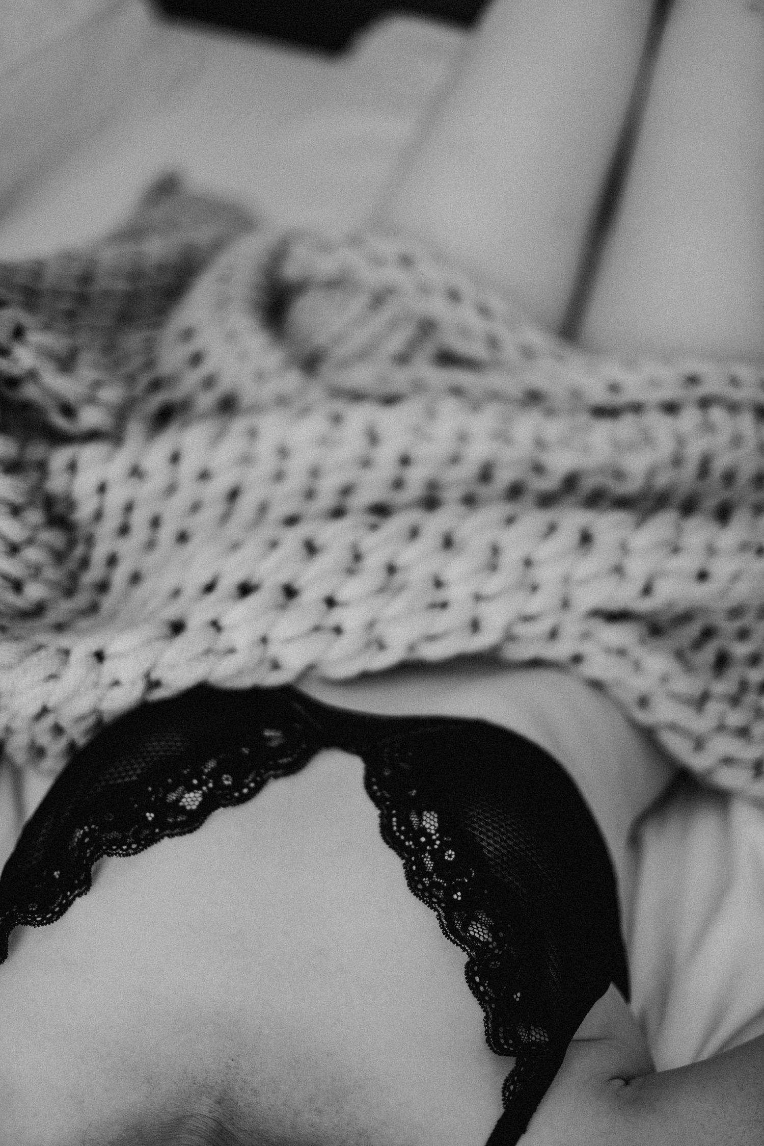 Person liegt auf einem Bett, teilweise mit einer gestrickten Decke bedeckt. Schwarze Unterwäsche ist sichtbar. Schwarz-Weiß-Foto.