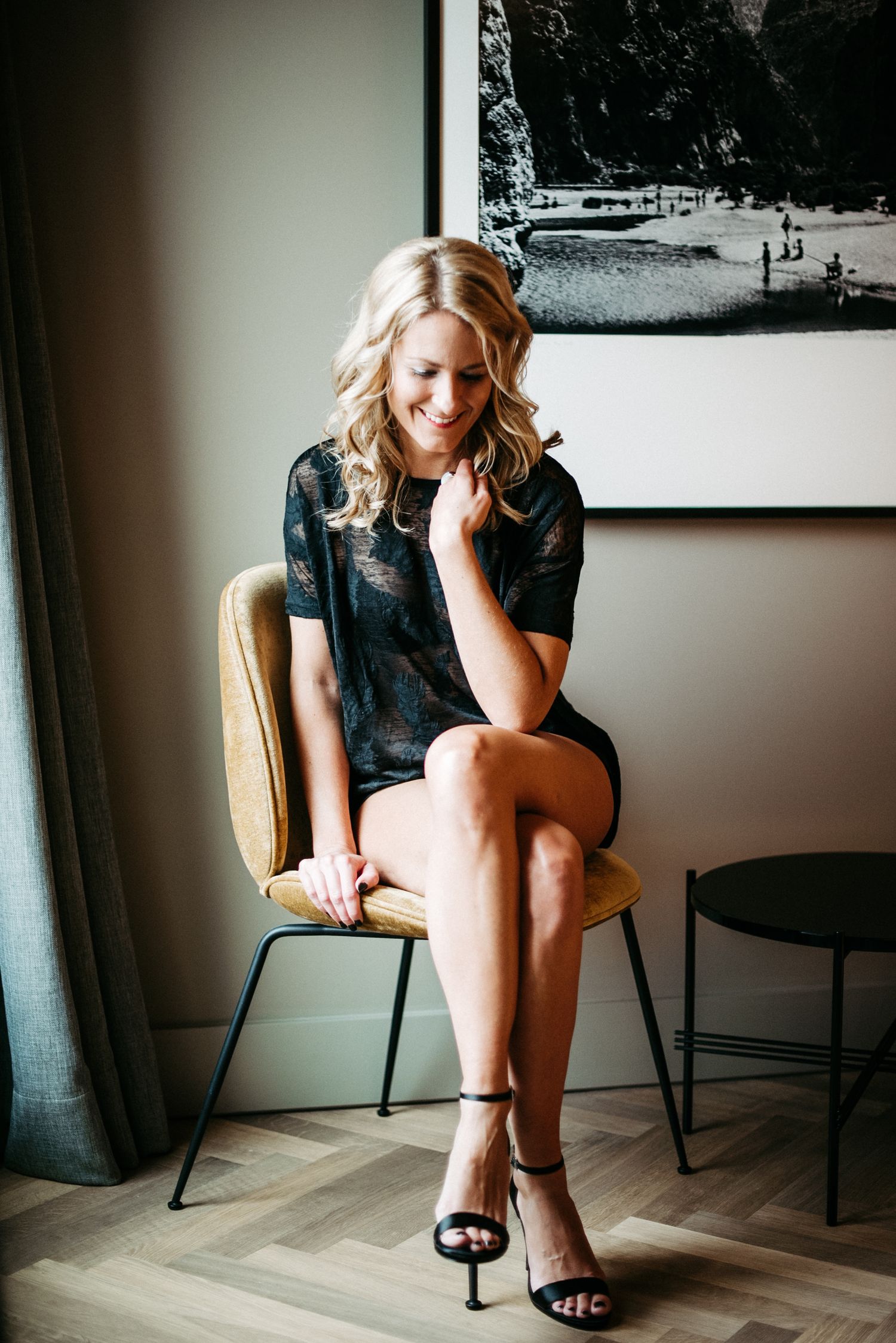 Frau in schwarzem Kleid sitzt lächelnd auf einem Stuhl in einem modern eingerichteten Raum mit einem großen gerahmten Foto an der Wand.
