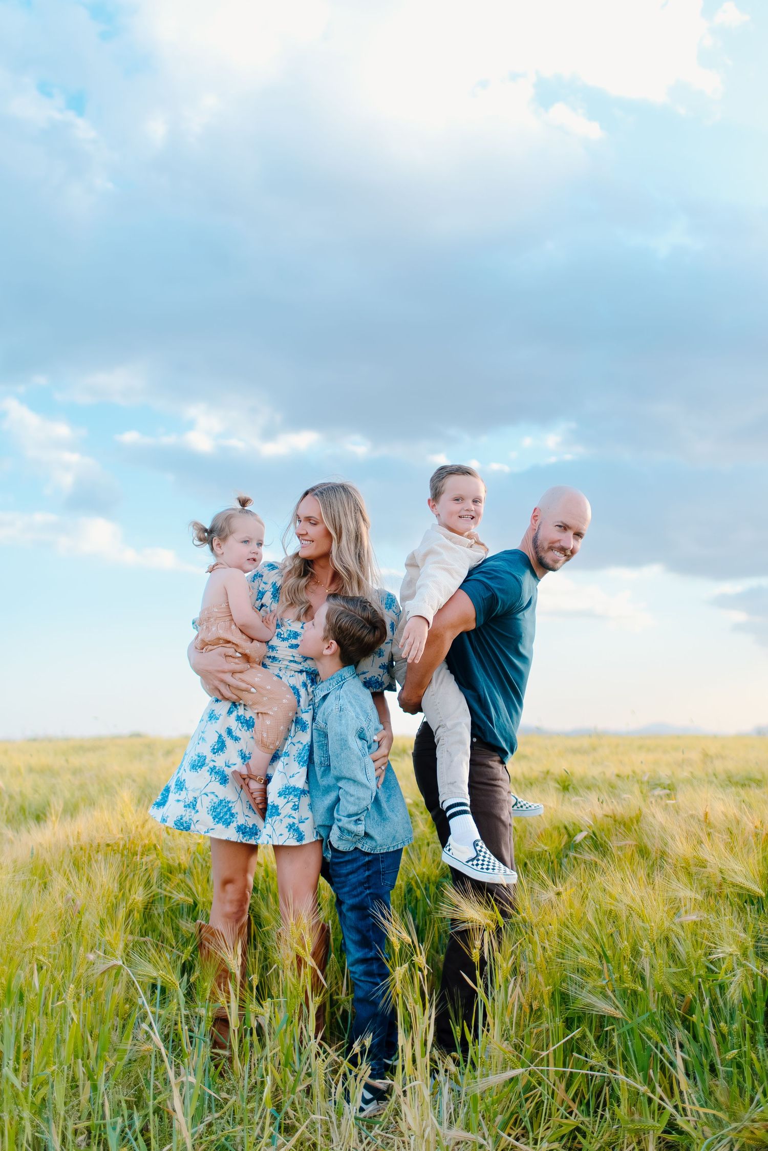 A family poses together in a sunny wheat field wearing coordinating blue outfits on a summer afternoon.