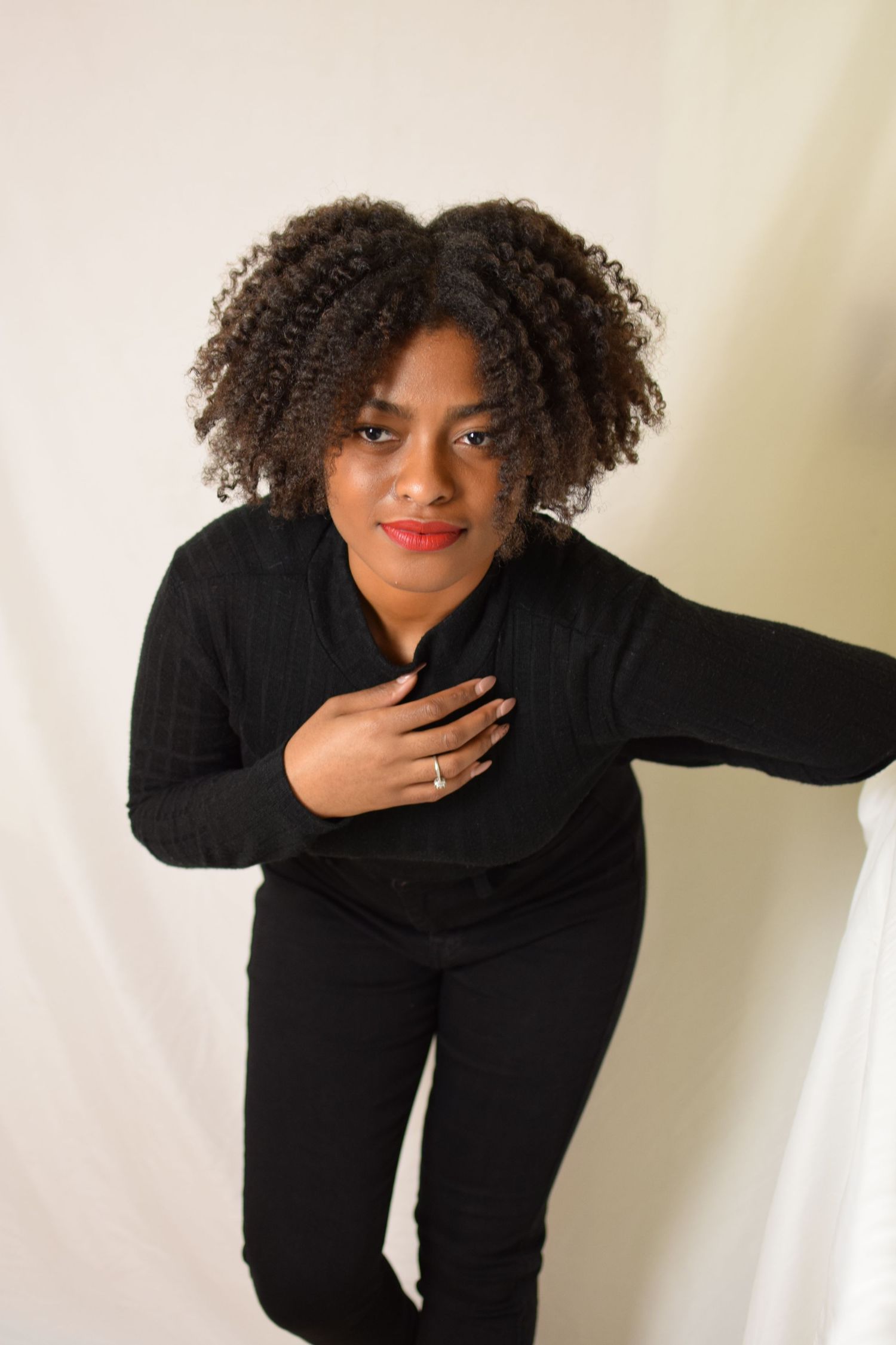 Person wearing all black outfit with natural curly hair poses against light background while leaning forward with hand on chest.