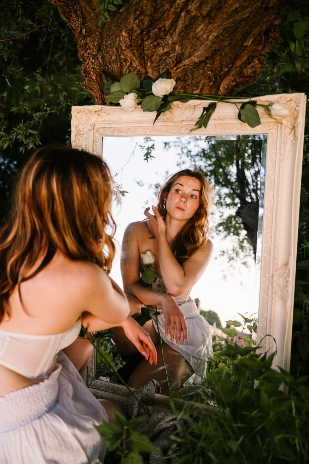 Artistic outdoor mirror reflection showing a person in a white dress crouching among green foliage and flowers.