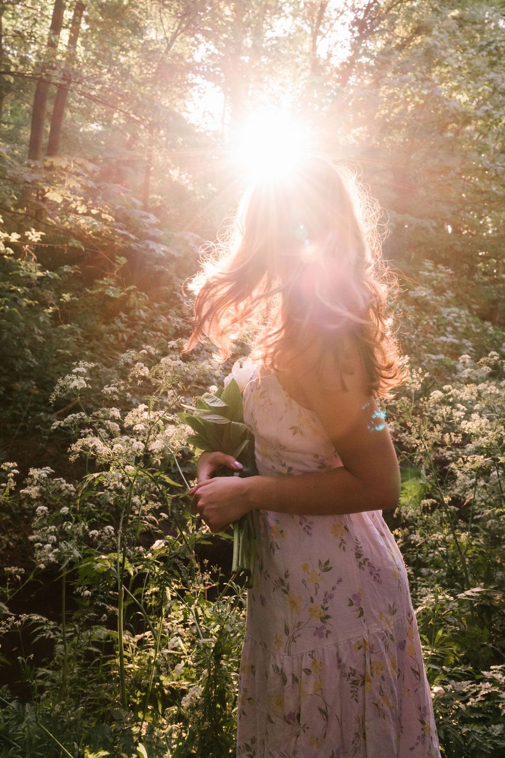 A person in a floral dress stands in a meadow of white wildflowers at sunset with sunlight creating a dreamy glow.