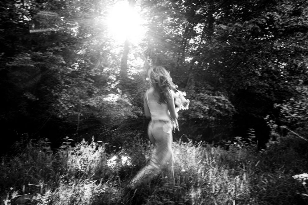 A dreamy black and white photograph captures a figure in nature reaching toward bright sunlight filtering through trees.