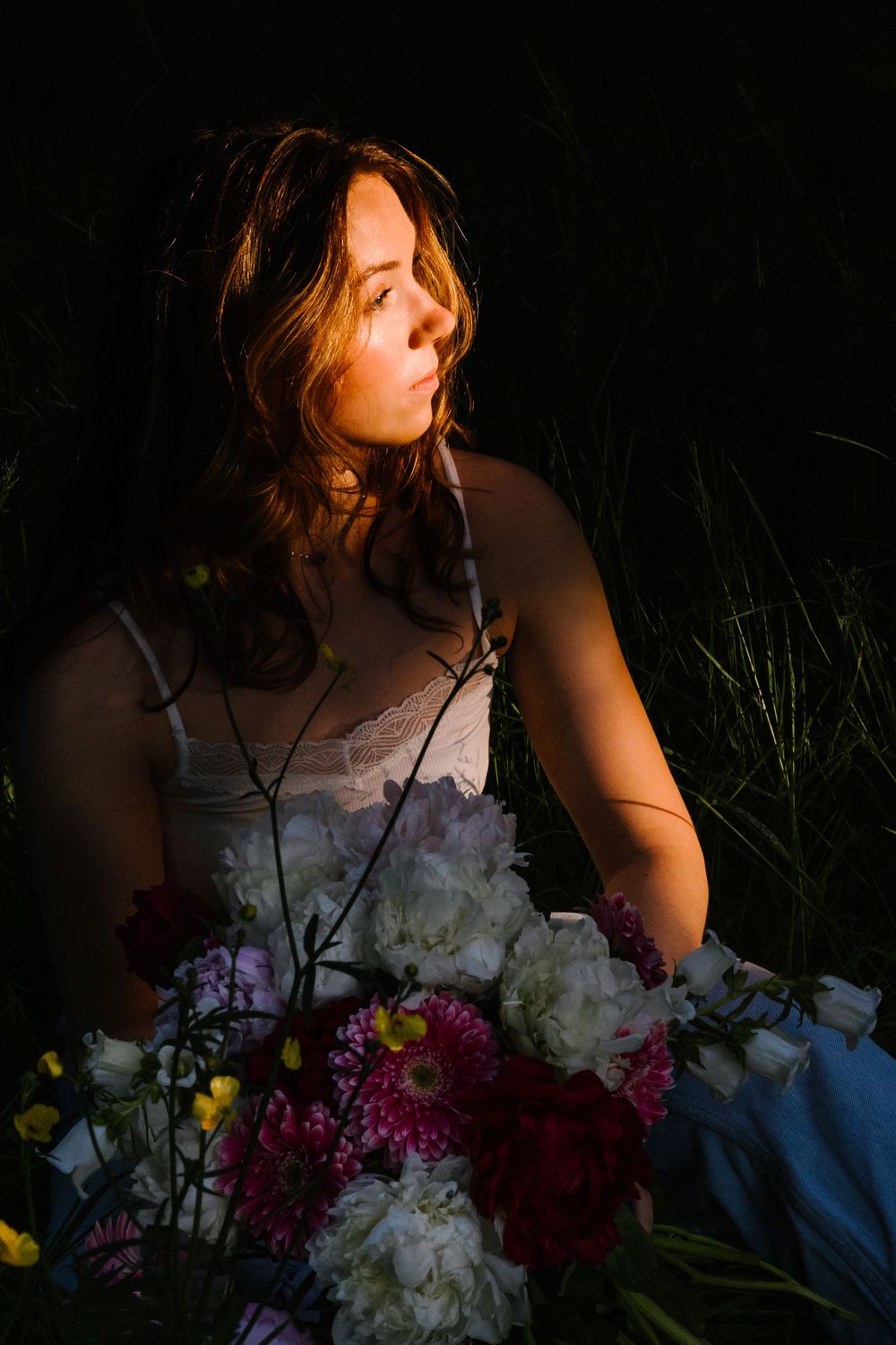 Moody portrait in darkness with dramatic lighting illuminating flowers and bare shoulders against black background.