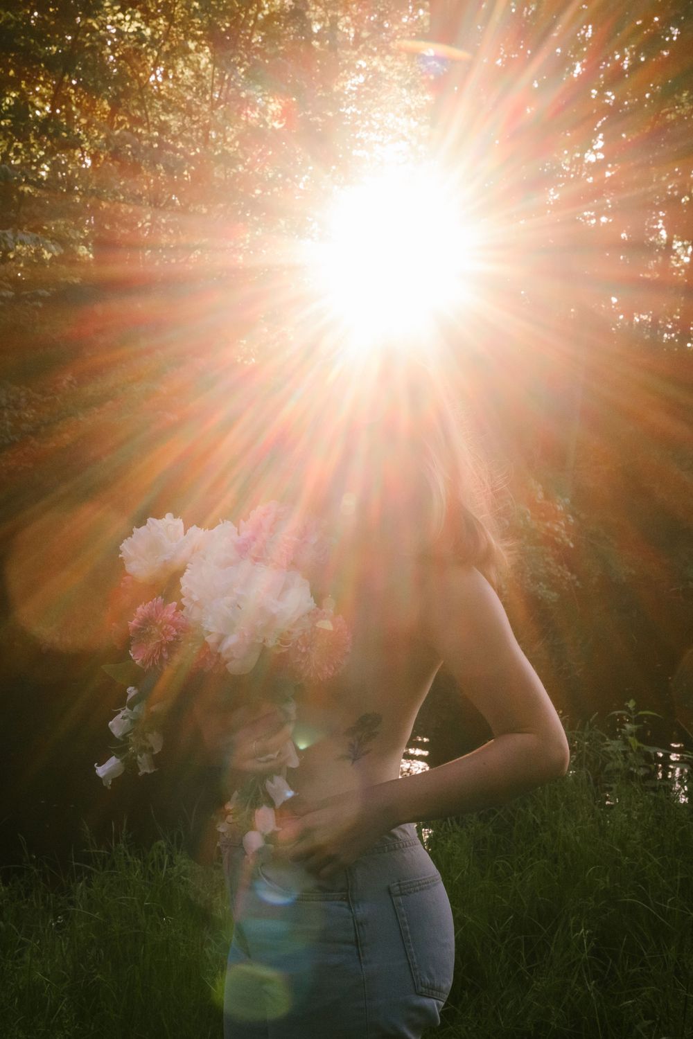 Bright sunlight bursts through trees creating dramatic lens flare behind silhouetted figure holding delicate white flowers.