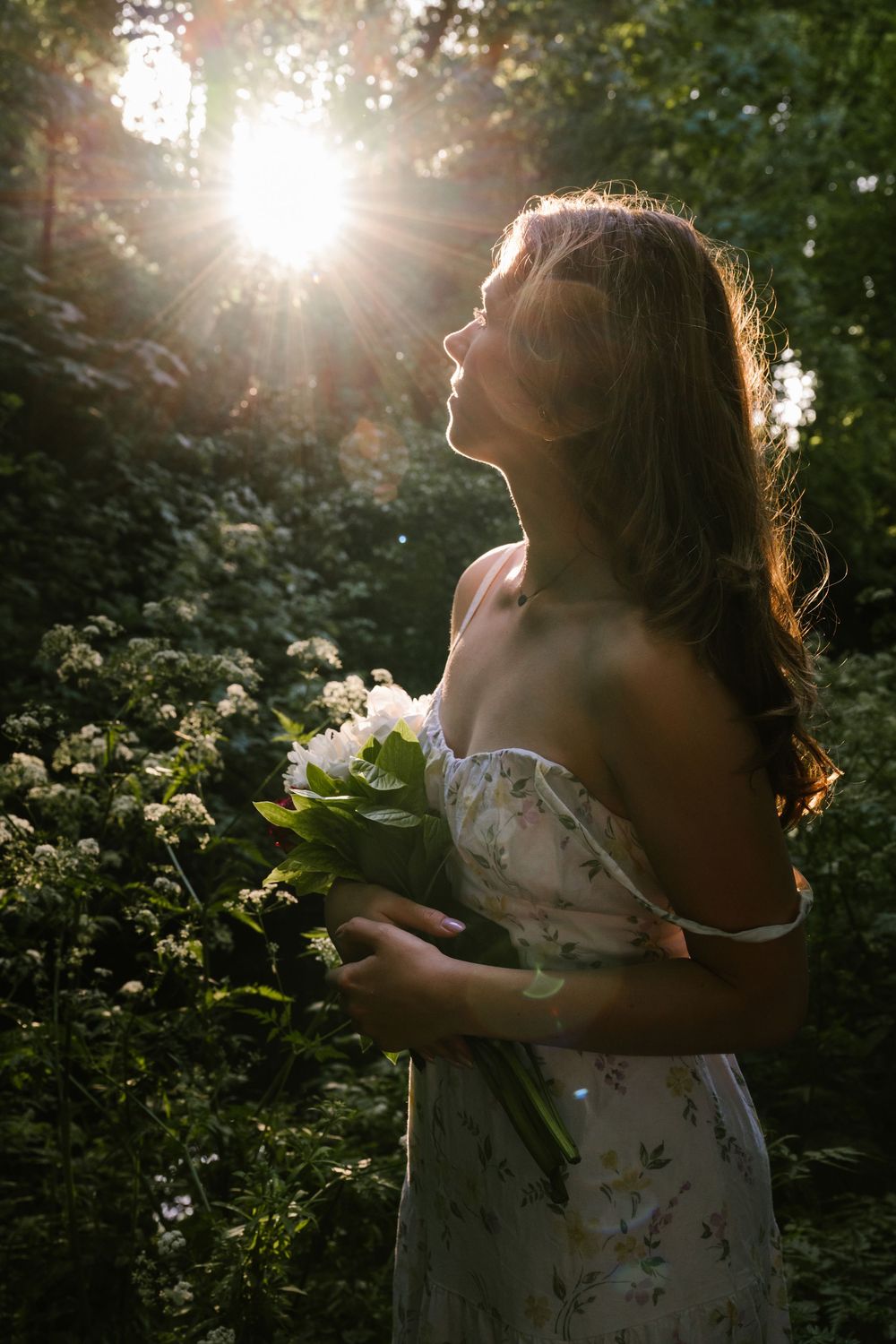 A figure in a floral dress stands in a forest with sunlight streaming through trees creating ethereal backlighting.