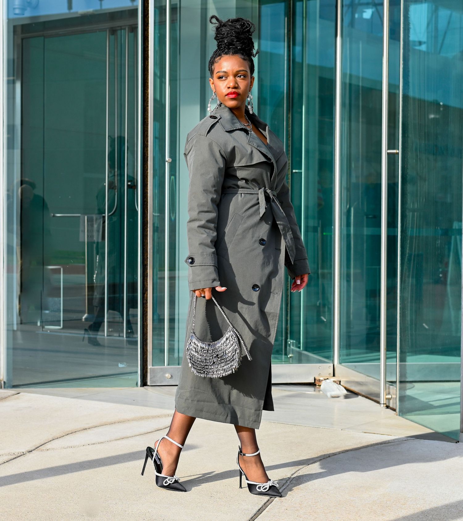 Stylish woman in grey trench coat and heels poses confidently outside modern glass building with silver handbag.