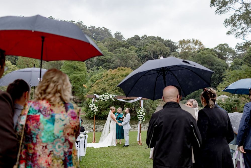 Wedding guests hold umbrellas during an outdoor garden ceremony on a rainy day.