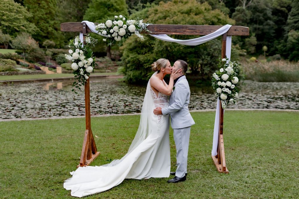 A couple shares their first kiss during an outdoor wedding ceremony under a wooden arch adorned with white flowers.