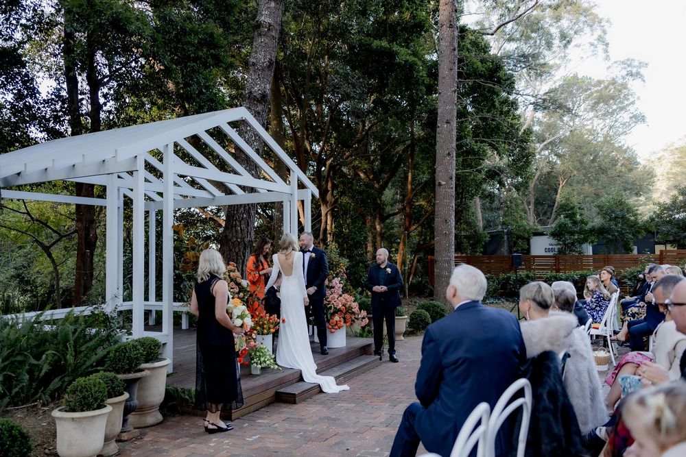 A beautiful garden wedding ceremony takes place under a white wooden pergola surrounded by trees and seated guests.