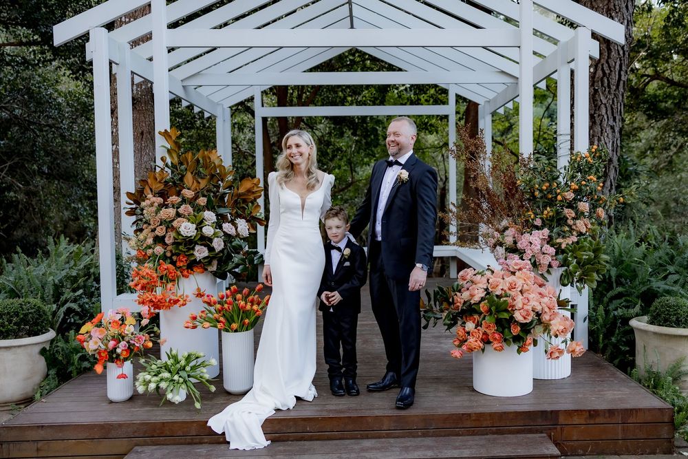Couple with their dog stands under a white gazebo decorated with vibrant orange and pink floral arrangements.