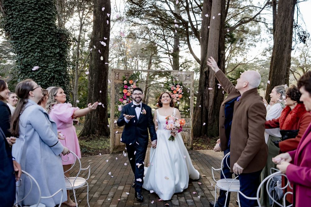 Happy wedding couple exits outdoor ceremony under evergreen trees while guests celebrate with pink ribbons.